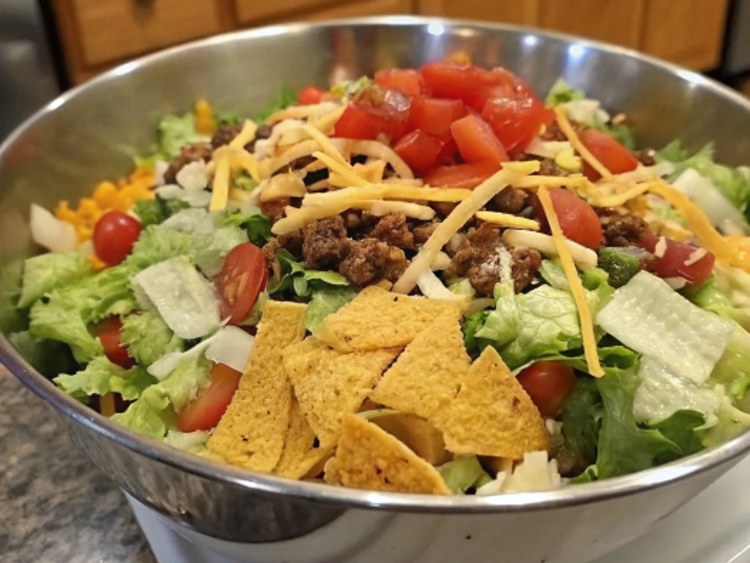 Ultra-close-up, high-quality image of a single Dorito Taco Salad, capturing its textures, colors, and details with intense focus. The salad is presented in a rustic ceramic bowl, tightly framed to showcase vibrant chopped tomatoes, shredded cheddar cheese, and flecks of red onion against a backdrop of golden, lightly crushed Doritos. Bright natural lighting highlights the glossy drizzle of ranch dressing and crispy edges of seasoned ground beef. Garnishes like fresh cilantro leaves and a lime wedge add a pop of color without overwhelming the dish. The background is softly blurred to emphasize the salad’s freshness and crunch.