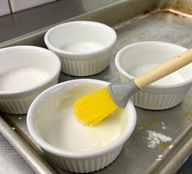 ultra-close-up view of a hand brushing butter inside a white ramekin, placed on a clean marble countertop. The scene is brightly lit to highlight the butter’s soft sheen and the smooth ceramic texture of the ramekin.