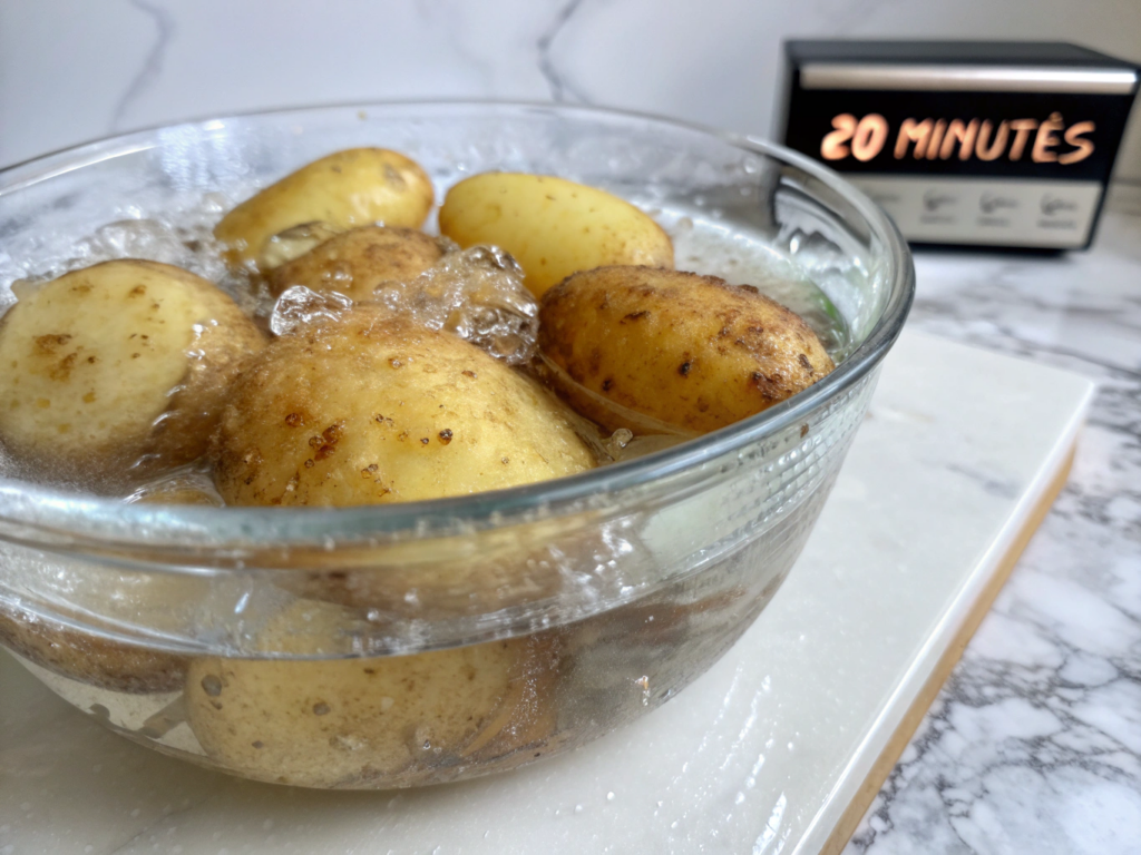 Ultra-close-up view of russet potato wedges submerged in a glass bowl of icy water, droplets clinging to their surfaces. Shot against a white marble countertop with a timer showing 20 minutes.